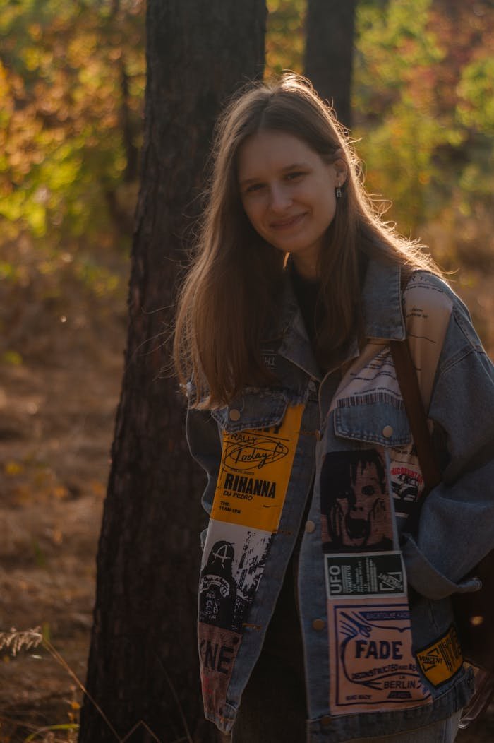 Young woman smiling in a park wearing a denim jacket, showcasing autumn fashion.