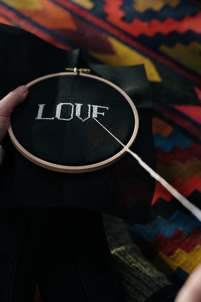 about-us-02 Close-up of hand embroidery hoop with 'Love' stitched on black fabric, colorful background.